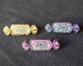 Three candy-shaped enamel pins on a dark textured background, featuring the text 'Chaotic Sweet' in white. One pin is gold, one is lavender, and one is pink, all with silver-toned metal edges.