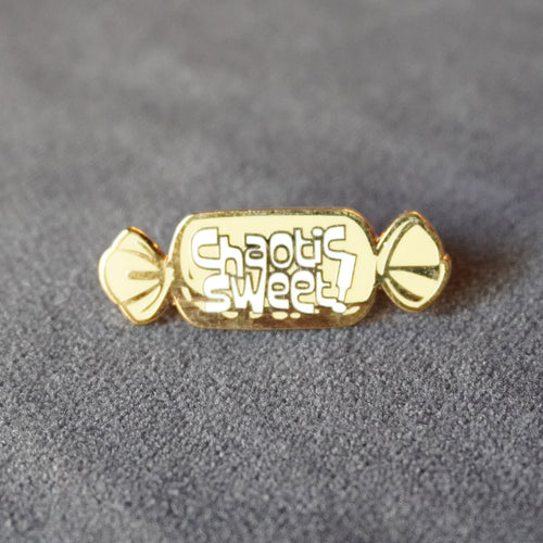 A gold candy-shaped enamel pin with the words 'Chaotic Sweet!' in white script, resting on a textured gray surface.