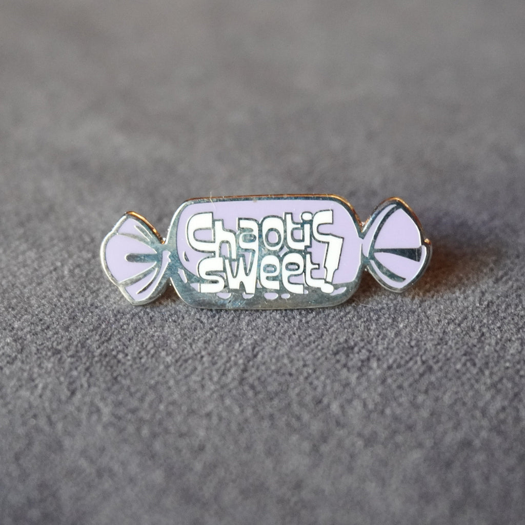 A candy-shaped enamel pin with the text 'Chaotic Sweet!' in white lettering on a light lavender background, with gold-toned metal outlines and silver-toned side details, resting on a textured gray surface.