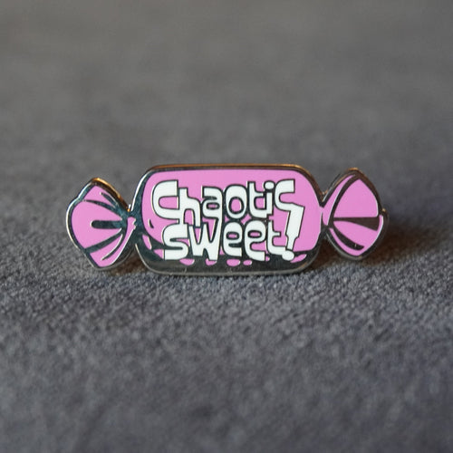A pink and silver enamel pin shaped like a wrapped candy, featuring the text 'Chaotic Sweet!' in white stylized lettering, resting on a textured gray surface.