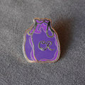 A small purple and lavender monogrammed bag-shaped pin with silver metal trim and an 'CR' monogram, resting on a textured gray surface.