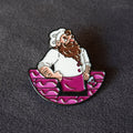 A cartoon enamel pin of a bearded chef in a white uniform sitting behind a donut wall photographed on a dark textured background.