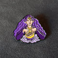 A small enamel pin shaped like a stylized woman with long flowing purple hair, seated over a yellow bowl, photographed on a dark textured surface.