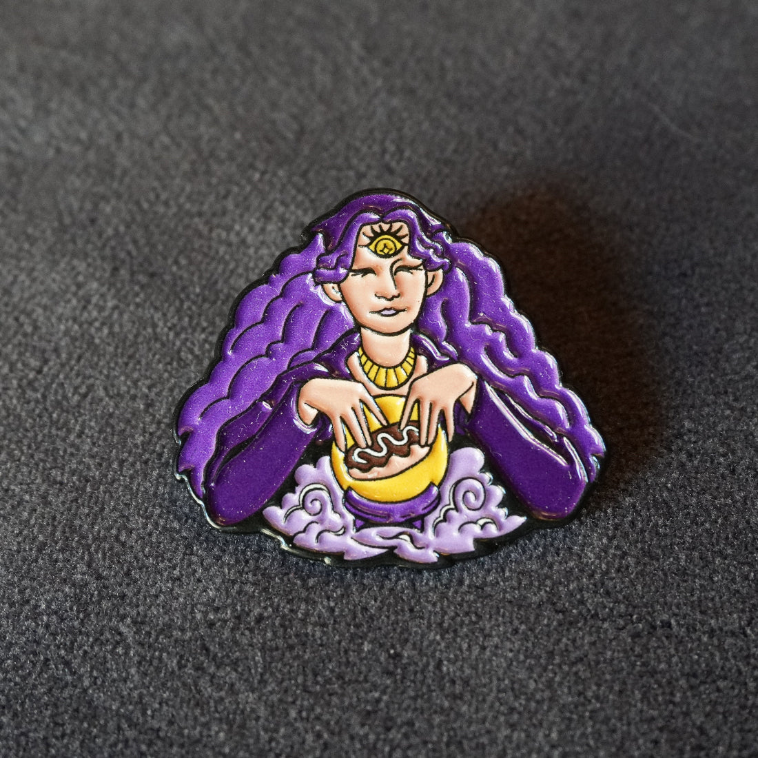 A small enamel pin shaped like a stylized woman with long flowing purple hair, seated over a yellow bowl, photographed on a dark textured surface.