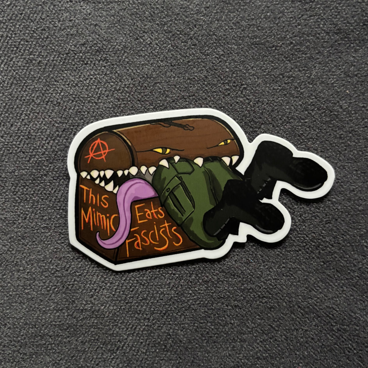A die-cut sticker featuring a cartoon mimic eating a fascist and the text 'This Mimic Eats Fascists' in orange, shown on a dark gray textured fabric background.