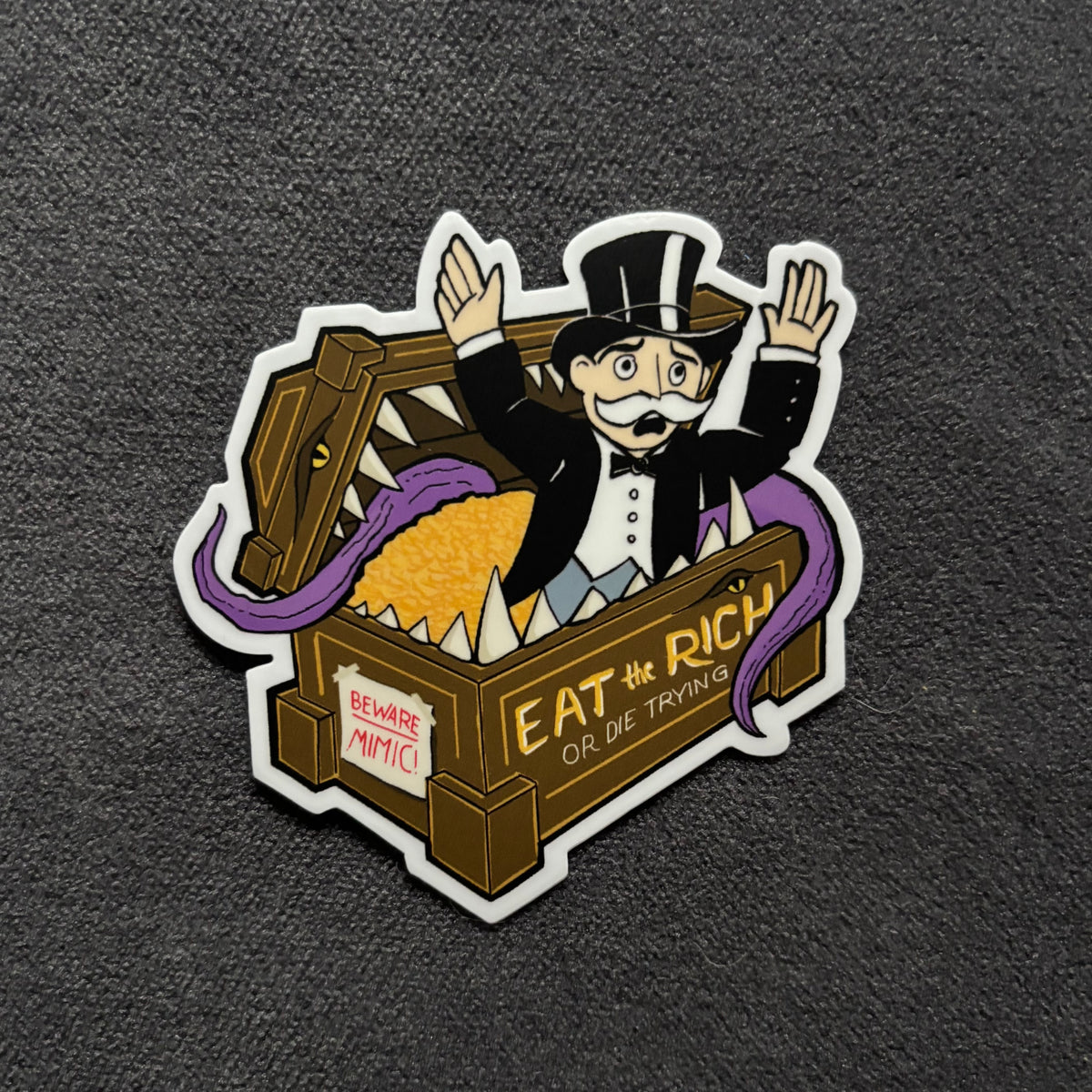 A die-cut enamel pin featuring a cartoon man in a top hat popping out of an open treasure chest filled with gold coins, with purple tongues. The chest has the text 'EAT THE RICH OR DIE TRYING' and a warning sign reading 'BEWARE MIMIC'.