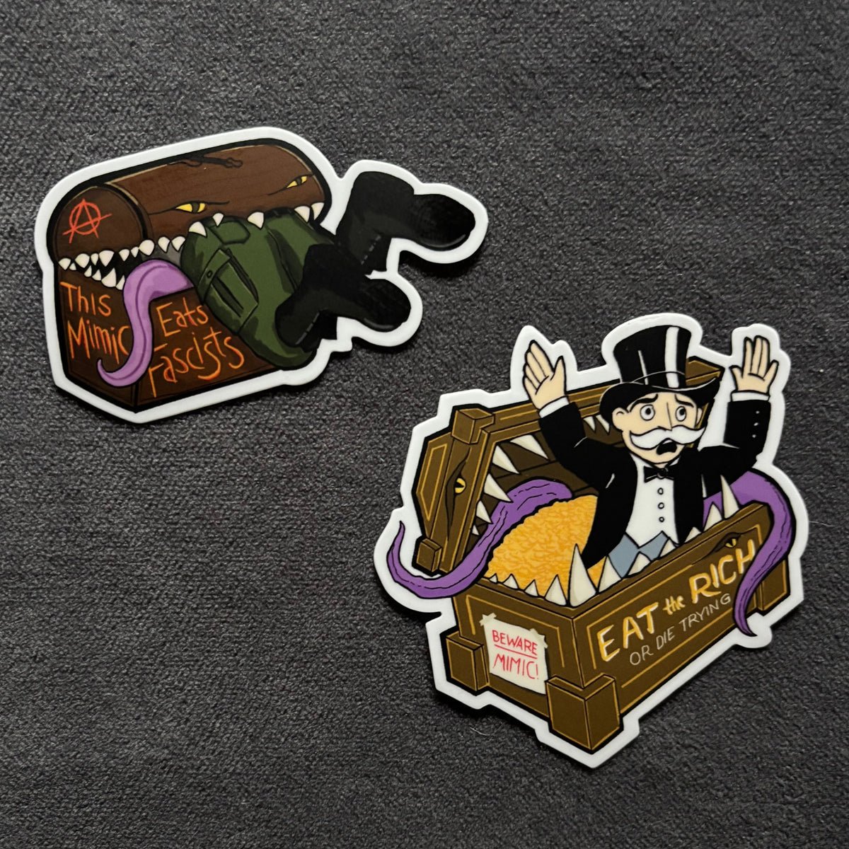Two cartoon-style die-cut stickers on a dark textured background. One sticker shows a fascist being eaten by a mimic and the other is a monopoly man being eaten. 