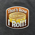 A die-cut sticker featuring a cinnamon roll illustration with the text 'That's How I Roll!' in bold white lettering, shown on a dark textured fabric background.