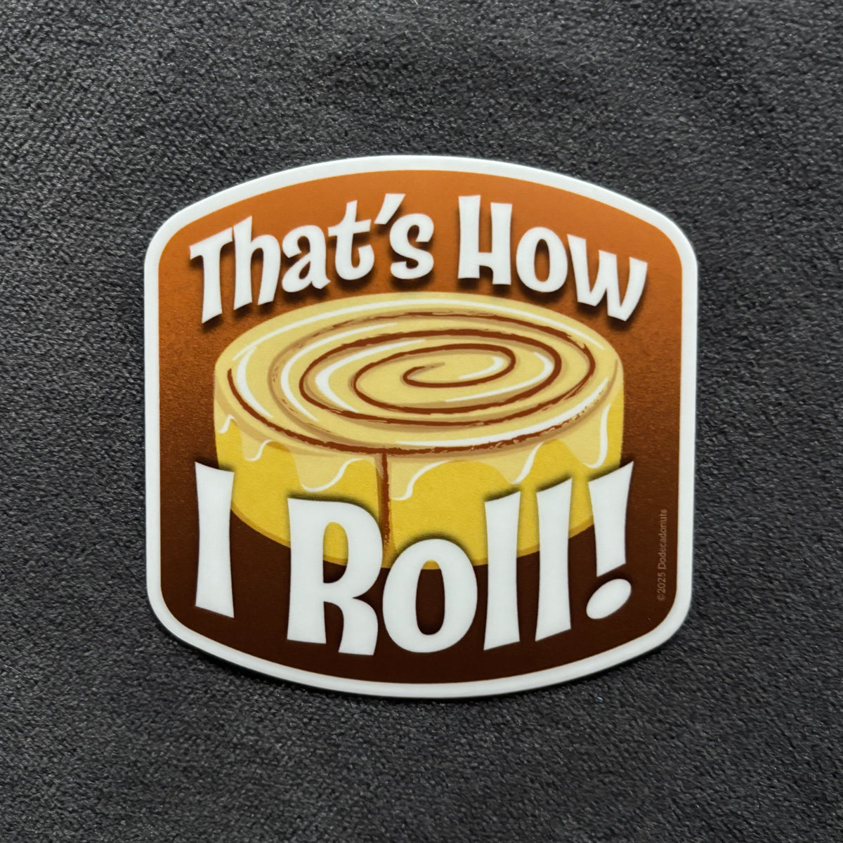 A die-cut sticker featuring a cinnamon roll illustration with the text 'That's How I Roll!' in bold white lettering, shown on a dark textured fabric background.