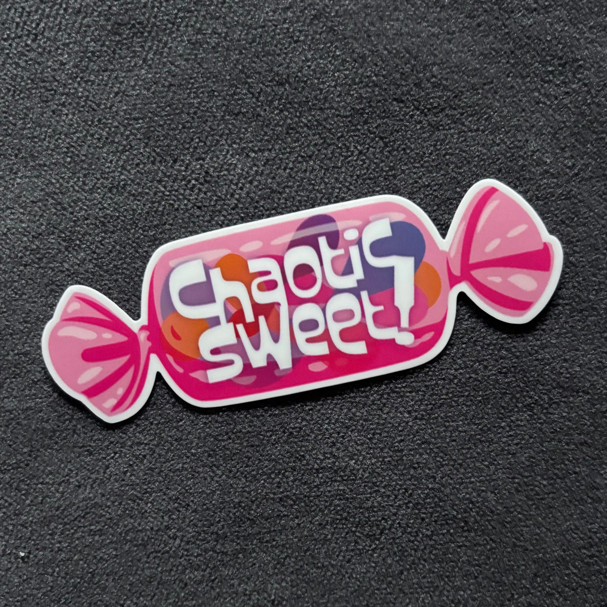 A die-cut sticker shaped like a pink candy wrapper with the words 'chaotic sweet!' in white lettering, placed on a dark textured background.