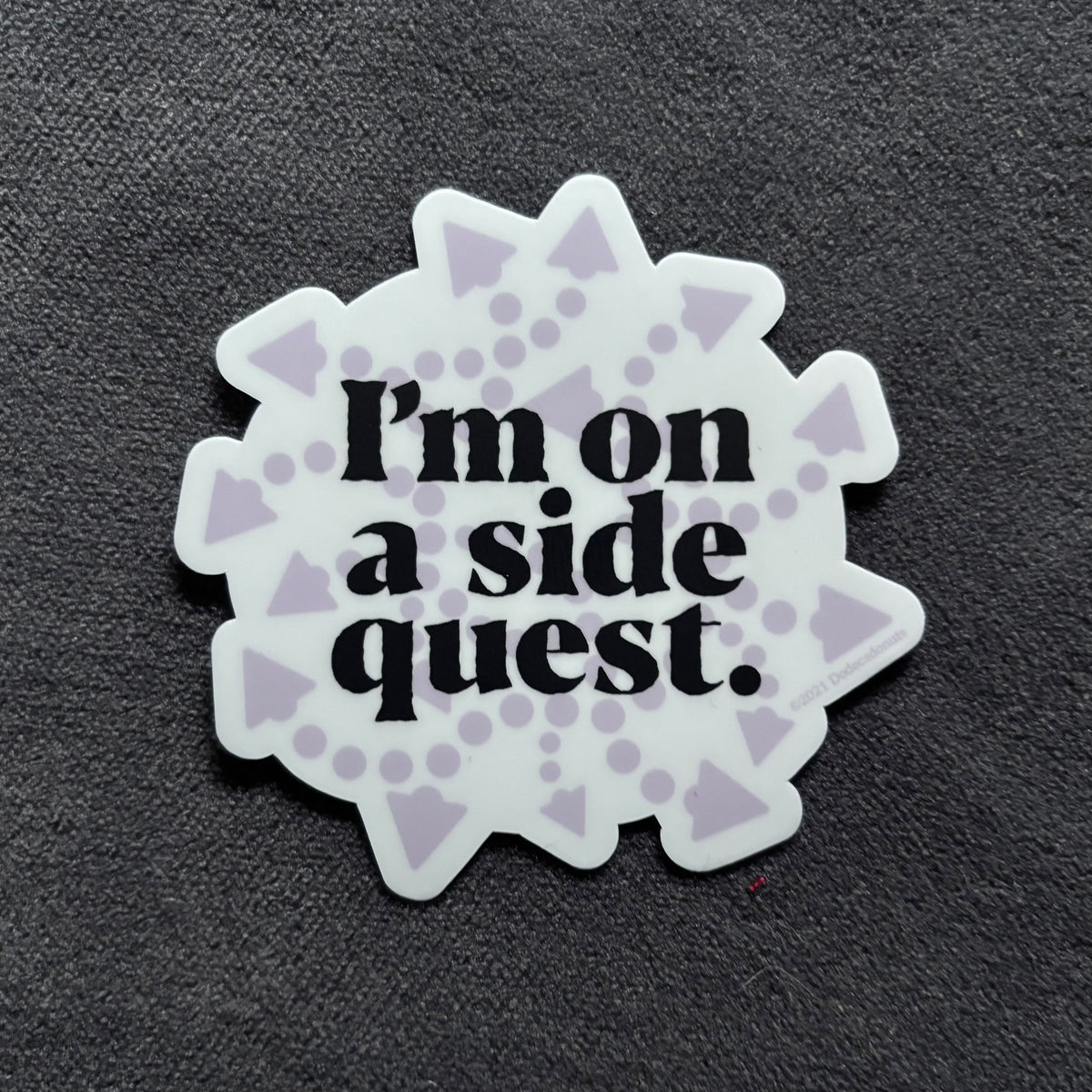 A die-cut sticker with the phrase 'I'm on a side quest.' in black serif text, surrounded by a white scalloped border with light purple geometric patterns, shown on a dark textured background.