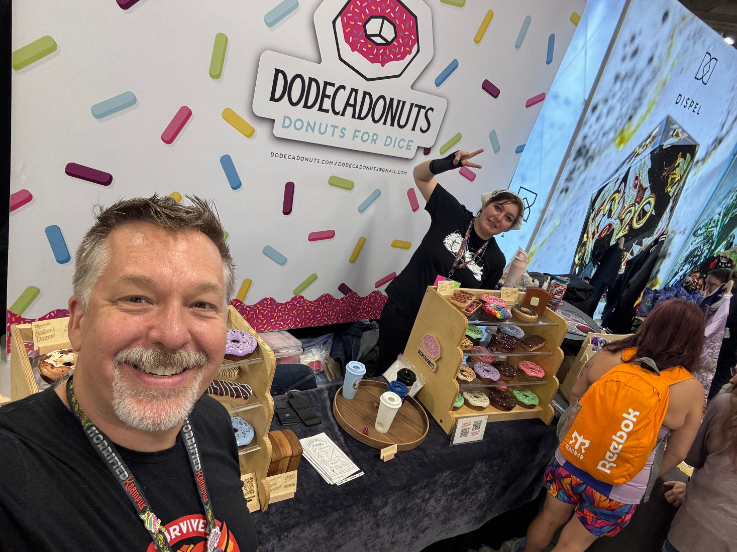 A busy convention booth for Dodecadonuts, with a smiling man taking a selfie in the foreground, a woman behind him gesturing toward the display. The booth features a white wall decorated with colorful donut-shaped sprinkles and a large Dodecadonuts sign.