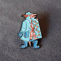 A small enamel pin shaped like a three kobolds wearing a light blue trench coat and blue boots, peeking out from behind the coat and orange cookies details on the coat. The pin is photographed on a dark gray textured surface.