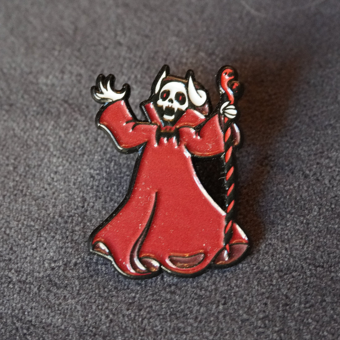 A gothic-style enamel pin depicting a skeletal figure in a flowing red robe holding a twisted staff, photographed on a textured dark gray background.