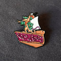 A small enamel pin shaped like a slice of pizza with a cartoon mouse dressed as a pirate standing on top, pointing forward, photographed on a dark textured surface.